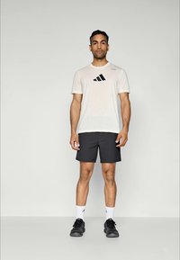 Lys beige trenings t-skjorte med svart Adidas-logo, kombinert med svarte shorts og svarte joggesko. Modellen står foran en ensfarget bakgrunn.