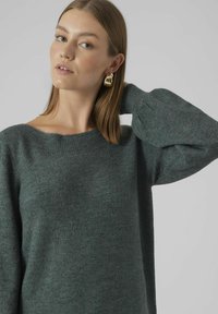Maglione verde lavorato a maglia con scollatura ampia e maniche a sbuffo. Tessuto morbido e vestibilità rilassata, accentuata dagli orecchini dorati della modella.