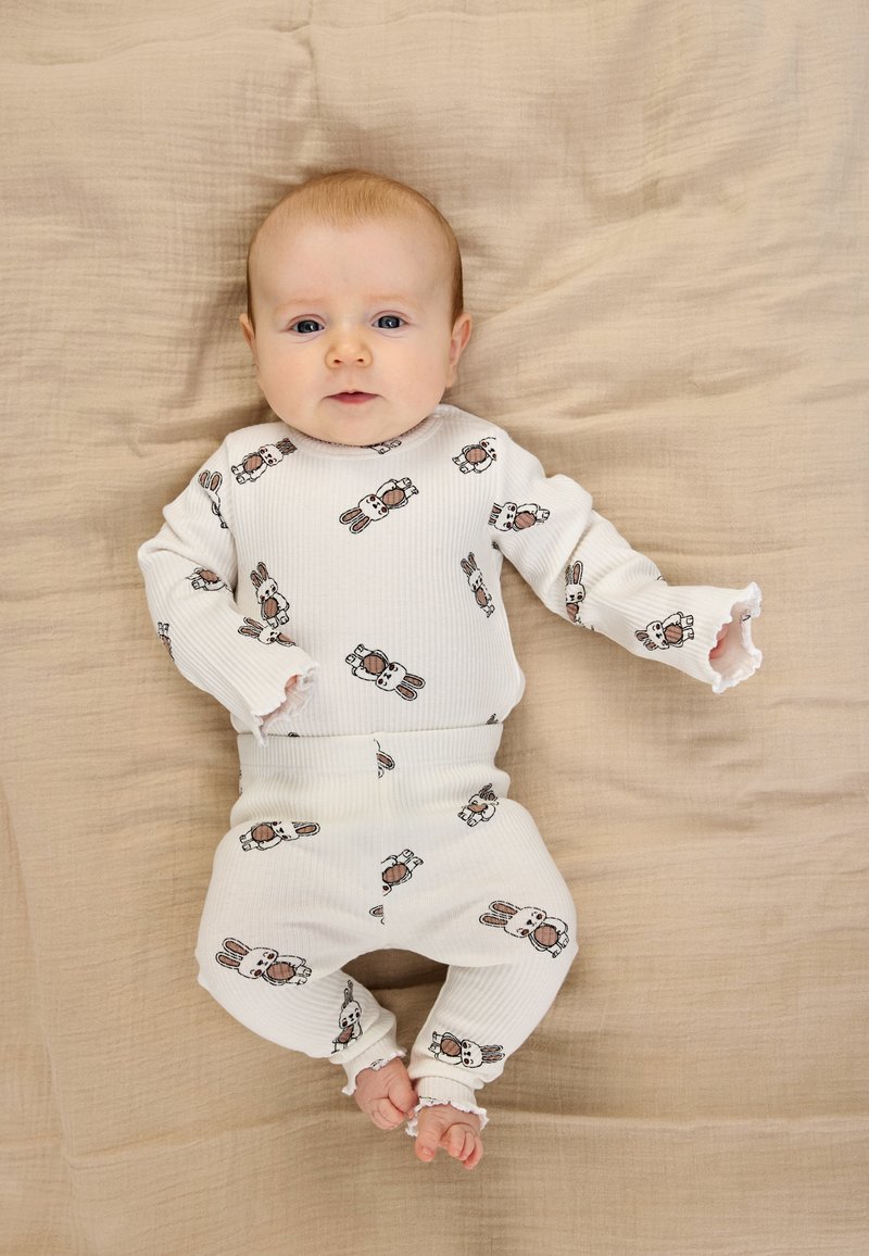 Hvid ribbet babyoutfit med lange ærmer og bukser, med et gentaget kaninmønster. Blød tekstur, flæsekant ved ærmer og ankler.