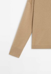 Manche et ourlet d'un pull en tricot beige à manches longues avec poignets et bord inférieur côtelés sur fond blanc.