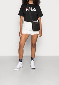 T-shirt Fila noire, shorts en denim blanc usé, sac bandoulière noir, baskets hautes blanches et chaussettes grises à motifs.