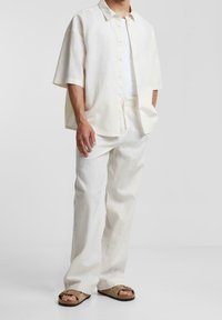 Camicia in lino a maniche corte color beige chiaro, abbottonata, abbinata a pantaloni larghi dello stesso colore e sandali marroni dalla texture morbida.