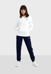 Sweatshirt à capuche blanche avec poche frontale, pantalon de survêtement bleu marine, baskets blanches, avec un petit logo sur la poitrine et sur le pantalon.