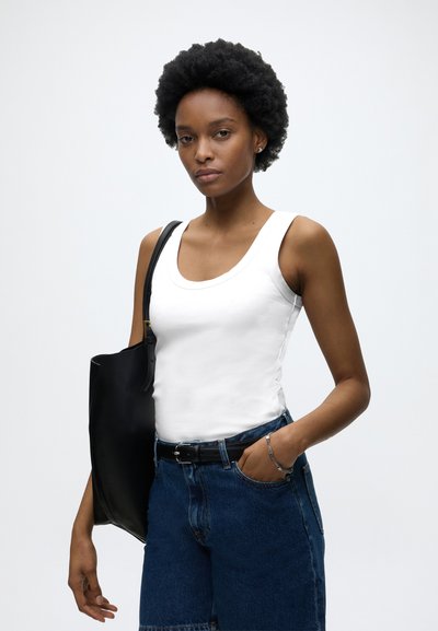 Γυναίκα με κοντά σγουρά μαλλιά, που φοράει λευκό αμάνικο τοπ, μπλε τζιν σορτς, μαύρη ζώνη και κρατάει μαύρη τσάντα ώμου.