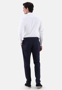 Camicia bianca a maniche lunghe con colletto alla francese, abbinata a pantaloni blu navy con passanti per cintura e due tasche posteriori, indossata da un modello visto da dietro.