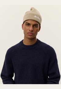 Beige knitted beanie with a pointed tip and white text detail, paired with a dark navy, chunky knit sweater featuring a round neckline.