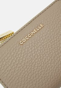 Bežinė odinė piniginė su tekstūruota paviršiumi, auksinės spalvos detalėmis ir įspaustu logotipu "Coccinelle" šone, su užtrauktuku.