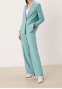 Blazer turquoise cintré et pantalon large en tissu lisse. Talons blancs complètent la tenue. Lignes épurées et coupe structurée.