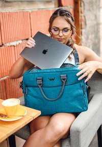 Türkise Laptoptasche mit grauen Akzenten, ausgestattet mit zwei Griffen und einem Reißverschluss, die mit einem silbernen Laptop gefüllt wird, neben einer gelben Kaffeetasse.