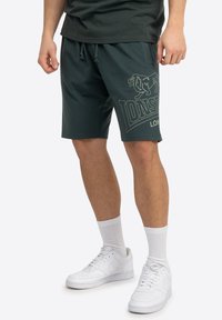 Grønne shorts lavet af bomuldsblanding, med et logo-print på siden og en elastisk taljebånd med snøre. Bæres med hvide sneakers og sokker.