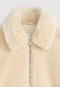 Krém színű műszőrme dzseki, amely széles gallérral, cipzáras záródással és puha, bolyhos textúrával rendelkezik, ideális a melegség és kényelem biztosításához.