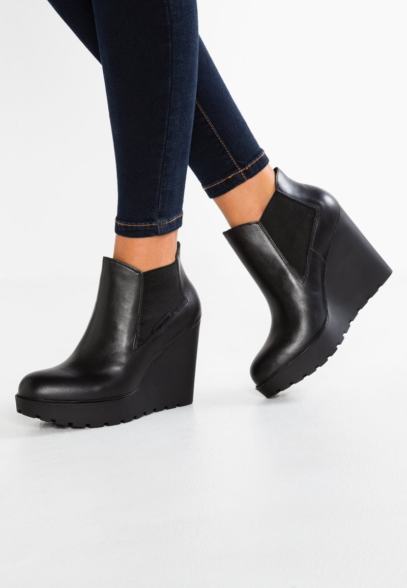 Botines negros de cuña con parte superior de cuero liso y paneles laterales elásticos. Detalles incluyen una suela texturizada y un diseño angular.