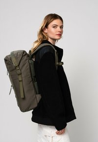 Sac à dos vert olive avec une surface texturée, des bretelles ajustables et une poignée supérieure, porté sur une veste en polaire noire.