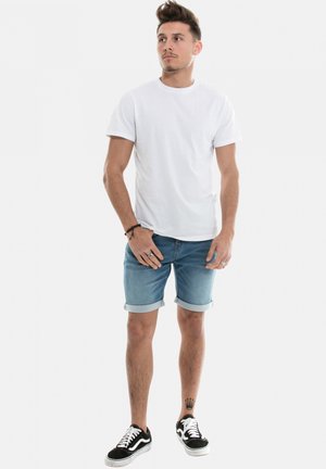 T-shirt en coton blanc, short en denim bleu clair avec ourlets retroussés, et baskets noires avec accents blancs. Figure masculine posant droite.