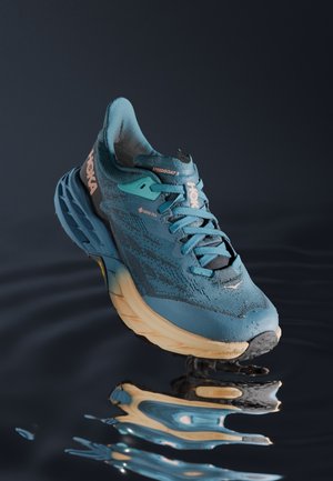 Niebiesko-beżowy but do biegania Hoka Speedgoat 5 z materiałem Gore-Tex odbijający się na ciemnej, falującej powierzchni wody.