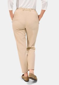 Pantalon beige avec une coupe ajustée, présentant une jambe droite et des passants de ceinture. Le tissu semble lisse avec deux poches arrière visibles.