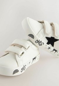 White sneakers with Velcro straps, black star accent, and playful black prints of a rainbow and animals on the sole. Textured leather material.