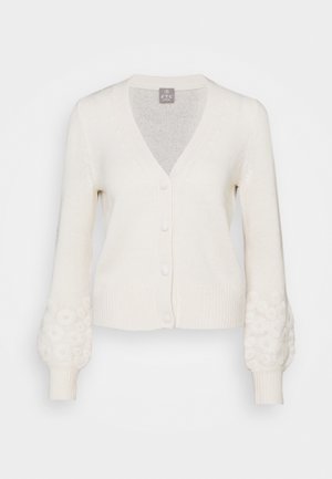 Gebreid vest in roomkleur met V-hals, knopen aan de voorkant, geribde zoom en manchetten, en een gestructureerd bloemmotief op de onderarmen.