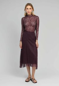 Haut en dentelle à manches longues de couleur violet profond avec un corsage ajusté. Jupe fluide de la même teinte, avec un ourlet transparent. Sandales noires avec des accents argentés.