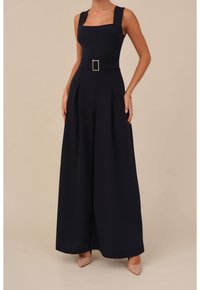 Mujer vestida con un mono negro de pierna ancha, con tirantes gruesos, escote cuadrado, un cinturón con hebilla de plata y tacones nude de punta.