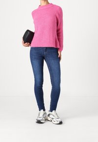 Pull en tricot rose avec une texture côtelée, associé à un jean skinny bleu. Porte des baskets argentées et tient une pochette noire.