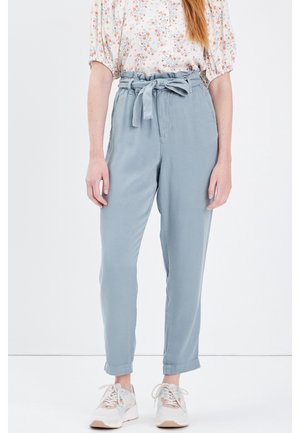 Vrouw die lichtblauwe broek met hoge taille en gestrikte ceintuur draagt, een gebloesde blouse met pofmouwen, en witte sneakers, staand tegen een witte achtergrond.