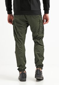 Olivgröna cargobyxor med elastiska muddar, flera sidofickor och en avslappnad passform, tillsammans med svarta sneakers på en vit bakgrund.