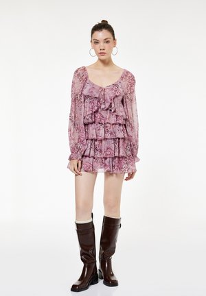 Nainen pukeutunut vaaleanpunaiseen kuviolliseen röyhelöiseen minimekkoon, jossa pitkät hihat, ja ruskeisiin polven korkeisiin saappaisiin, seisoo yksivärisen taustan edessä.