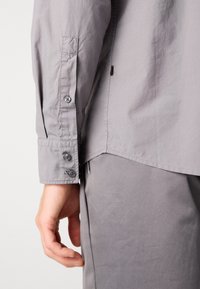 Camicia grigia con bottoni, dal tessuto liscio, con maniche lunghe, tre bottoni ai polsini e un orlo leggermente curvo.