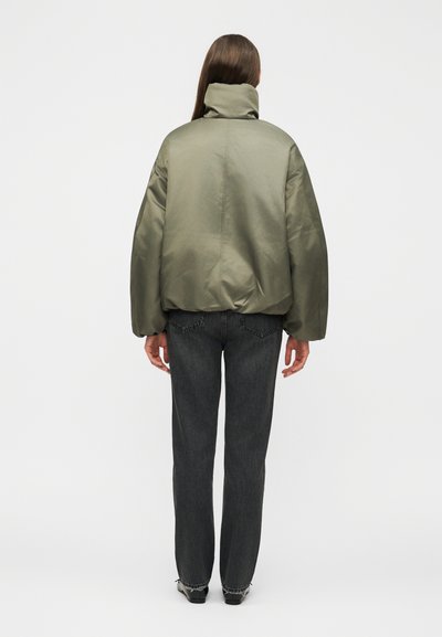 Άτομο με μακριά μαλλιά που φοράει ένα χακί φουσκωτό μπουφάν με ψηλό γιακά και μαύρα τζιν με ίσια μπατζάκια, στέκεται με την πλάτη προς τα εμπρός.