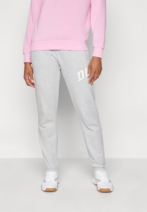 Graue Jogginghose aus weichem Material, mit einem weißen Logo auf dem linken Oberschenkel, kombiniert mit einem hellrosa Sweatshirt und weißen Sneakers.