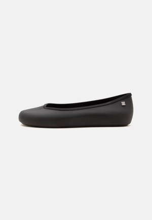 Chaussure ballerine plate noire avec bout rond et petit logo argenté sur le côté du talon, présentée sur fond blanc uni.