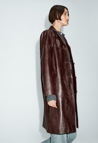 Cappotto in pelle bordeaux caratterizzato da un design lungo e strutturato, con bottoni frontali, due tasche laterali e dettagli di cucitura in evidenza.