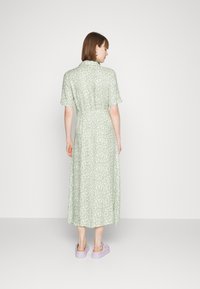 Vestido verde de comprimento até ao joelho com padrão floral, mangas curtas, tecido suave, cintura ajustada e sapatos lilás. Visto de costas.