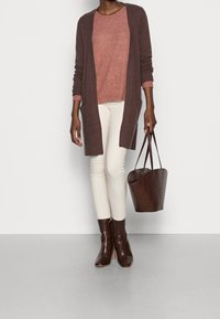 Lang brun cardigan over en lyserød top, parret med cremefarvede bukser og brune ankelstøvler i krokodilleskind. Holder en matchende brun tote-bag.