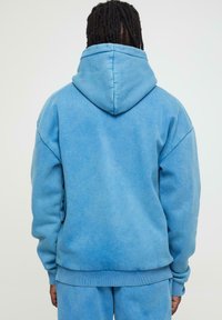Sweatshirt à capuche bleu clair, fabriqué en tissu doux avec une coupe décontractée. Présente des poignets et un ourlet côtelés, avec une surface lisse et légèrement texturée.