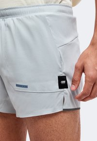 Gros plan sur un short de sport gris clair avec taille élastique, poche latérale, détail réfléchissant et écusson logo noir Ciele Athletics près de la cuisse.