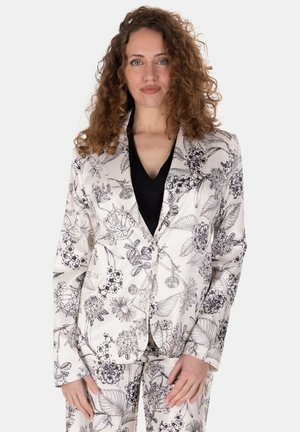 Donna con capelli ricci che indossa un completo bianco con stampa floreale nera e una maglia nera con scollo a V, in piedi davanti a uno sfondo chiaro e uniforme.