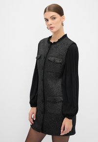 Vestido de cambio negro y gris con tejido texturizado, escote con volantes, cierres de botón y mangas negras con plisado. Presenta dos bolsillos en el pecho.
