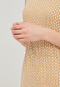 Beigių rankovių oranžinė suknelė su baltais ir žaliais taškeliais, turinti raukinį juosmens srityje ir tekstūruotą audinį. Rankos laiko audinį šalia juosmens.