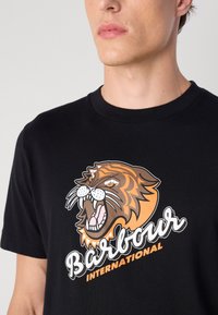 Tricou din bumbac negru, având un grafism cu un cap de tigru fioros în nuanțe de portocaliu și maro, însoțit de textul "Barbour International" în alb.