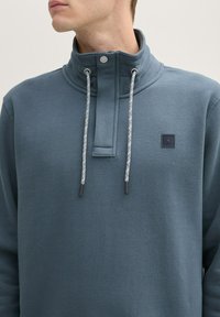 Sweat-shirt bleu foncé avec un col haut, des cordons gris et une finition texturée. Il comporte une petite étiquette logo sur la poitrine.