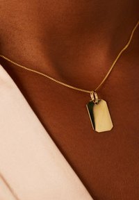 Collier en or avec un pendentif rectangulaire lisse, ayant un éclat subtil, sur un fond de peau avec un doux tissu drapé à proximité.
