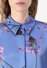 Ανοιχτό μπλε πουκάμισο με βοτανικό σχέδιο που περιλαμβάνει ροζ και μωβ λουλούδια. Σχεδίαση με γιακά, λεία υφή και κουμπί κλεισίματος στον λαιμό.