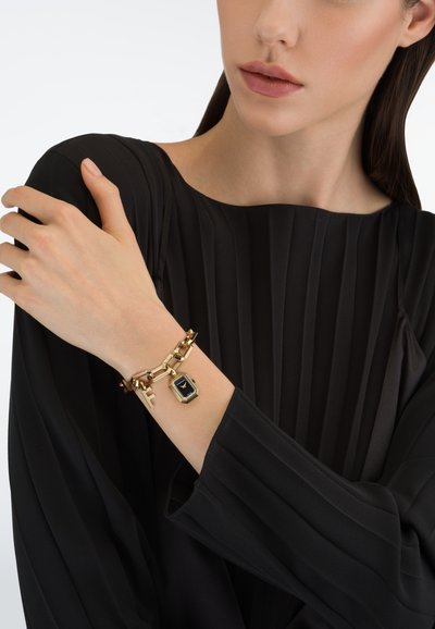 Gouden armband met een zeshoekige wijzerplaat en geometrisch schakeldesign, met een zwarte wijzerplaat. Model draagt een geplooide zwarte top.