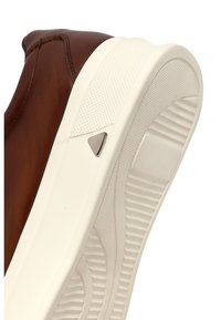 Sneaker in pelle marrone con suola in gomma bianca testurizzata, dotata di un accento metallico triangolare vicino al tallone. Parte superiore liscia con dettagli di cucitura.