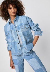 Denim-takki, jossa on patchwork-kuvio vaaleansinisissä sävyissä, kahdella etutaskulla, napitus ja käännetyt hihat.