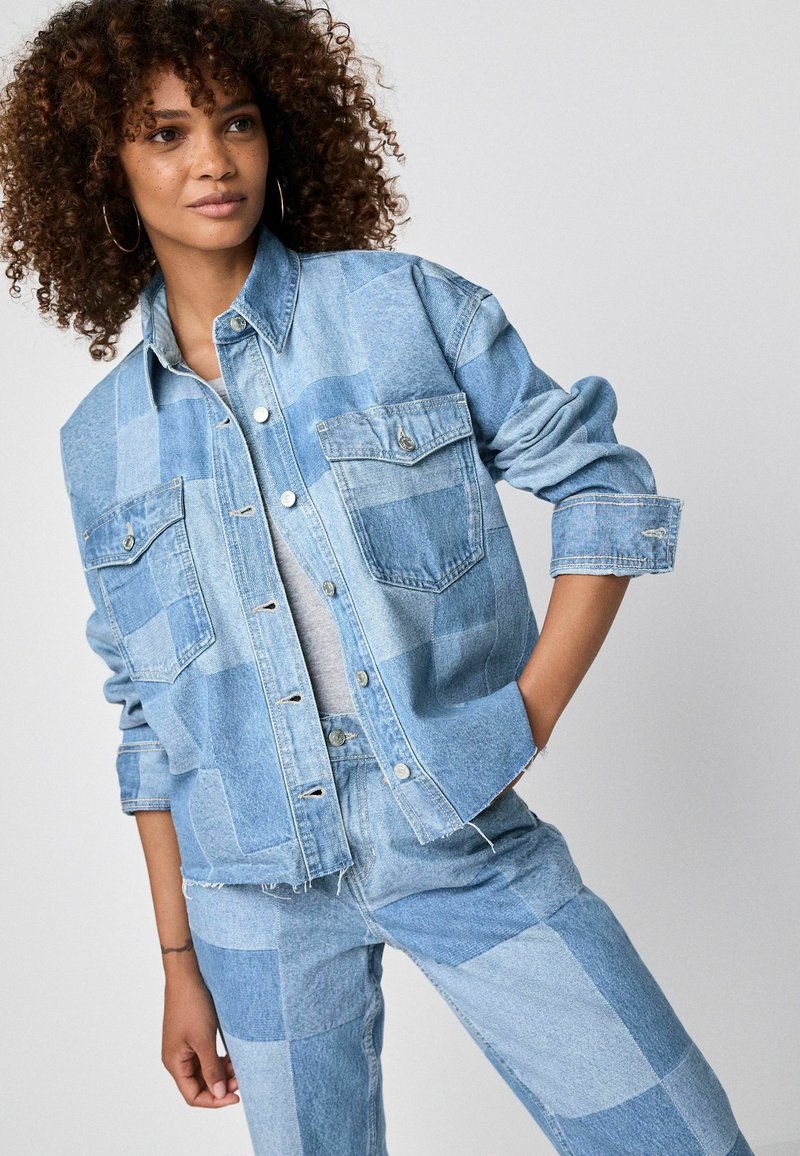 Denim-takki, jossa on patchwork-kuvio vaaleansinisissä sävyissä, kahdella etutaskulla, napitus ja käännetyt hihat.