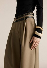 Un pantalon large beige avec des plis, associé à un haut noir à manches longues côtelé et des ceintures noires superposées. Des bracelets texturés en or au poignet.