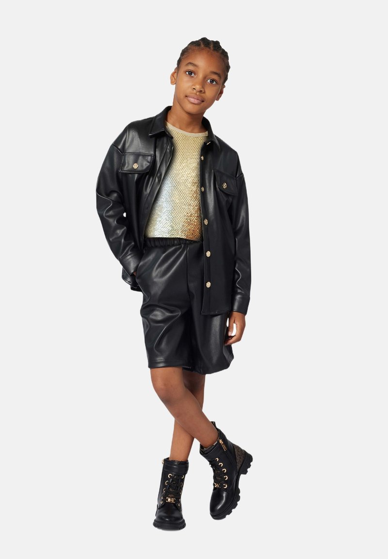 Veste oversize en simili cuir noir, haut métallique doré, short noir et bottes de combat noires avec des éléments en or. Le modèle se tient devant un fond blanc.